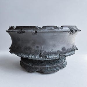 Pode incluir: Uma taça cinzenta escura com rebordo largo e superfície texturizada e desgastada. O exterior apresenta um rebordo alargado e recortado e uma base espessa e estriada. O acabamento mostra sombras manchadas e padrões finos e rachados.