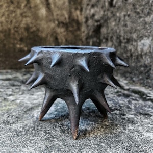 Pode incluir: Uma tigela de cerâmica cinza escuro, artesanal, com uma superfície texturizada e uma série de pontas cônicas e afiadas ao redor da borda. A tigela é elevada sobre três pernas pontiagudas, criando uma aparência escultural única.