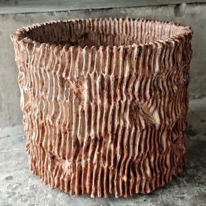 Pode incluir: Um vaso cilíndrico com uma textura rugosa que lembra a casca de uma árvore. O vaso castanho-avermelhado apresenta ranhuras verticais e uma borda irregular. O design evoca um estilo natural e rústico, adequado para uso interno ou externo.
