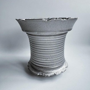 May include: A grey ceramic vase with a textured surface, featuring a wide, scalloped rim and a ribbed, cylindrical body. The vase has a flared base and a slightly rough, aged appearance, suggesting a handcrafted or vintage item.