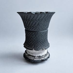 May include: A black vase with a flared rim and a textured, herringbone-like pattern covering the upper and middle sections. The base features a contrasting white, speckled finish that transitions into the dark, matte black body of the vessel.