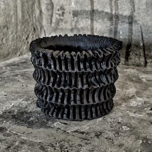 May include: A black, textured, cylindrical planter with a layered, ruffled design. The dark, matte finish and unique texture create a modern, artistic aesthetic. Ideal for small plants or as a decorative accent piece.