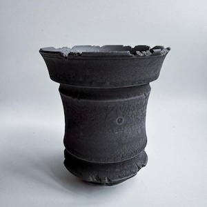 May include: A dark gray, textured ceramic pot with a unique, layered design. The pot features a wide, slightly jagged rim and a textured surface, giving it a rustic appearance. The pot's shape is reminiscent of a classical column.