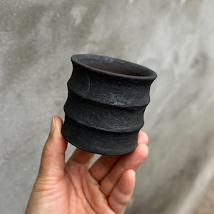 Puede incluir: Una pequeña maceta de cerámica negra con un exterior texturizado y acanalado. La maceta tiene una parte superior ancha y abierta y un interior oscuro. La cerámica hecha a mano se sostiene en una mano sobre un fondo gris neutro. La maceta probablemente se usa para plantas o como artículo decorativo.