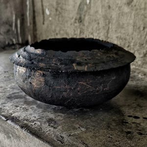 May include: A dark, textured bowl with a wide, uneven rim. The bowl is a deep black color, with visible texture on the surface. The interior is a dark void. The bowl sits on a concrete surface.