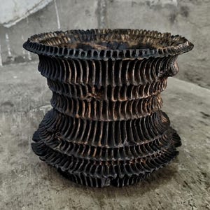 May include: A dark brown, textured, handmade ceramic vase with a unique, layered design. The vase has a wide, ruffled top and a flared base, with vertical grooves creating a ribbed effect. The overall aesthetic is rustic and artistic.