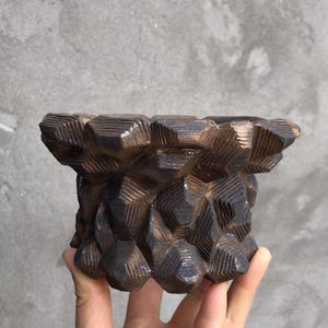 Puede incluir: Una maceta de cerámica marrón oscuro con un patrón geométrico. La maceta tiene una superficie texturizada con una serie de facetas angulares en forma de diamante. La maceta se sostiene en una mano sobre un fondo gris.