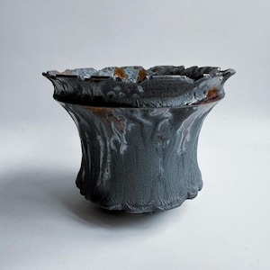 Puede incluir: Un jarrón de cerámica gris oscuro con una superficie texturizada y un borde rizado y acampanado. El borde presenta detalles en marrón y naranja. El jarrón tiene una base acampanada, creando una forma artística distintiva.