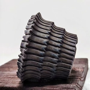 Puede incluir: Una escultura abstracta oscura y texturizada con un diseño en forma de ola. El objeto está hecho de un material oscuro, posiblemente metálico, y se encuentra sobre una superficie de madera. La forma de la escultura es compleja, con múltiples capas y crestas.