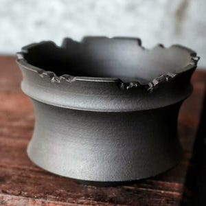 Op de afbeelding: Een donkergrijze keramische kom met een unieke, getextureerde rand. De kom heeft een brede, gebogen basis en een licht uitlopende bovenkant. Het oppervlak lijkt mat en het algehele ontwerp is minimalistisch en modern. De kom is waarschijnlijk handgemaakt.