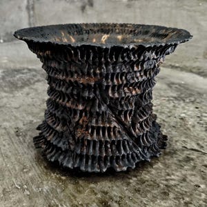 Puede incluir: Un jarrón artesanal marrón oscuro texturizado con un borde ancho y ligeramente ensanchado. El jarrón tiene un diseño único en capas con una apariencia rugosa y orgánica. El exterior es de un marrón profundo y carbonizado.