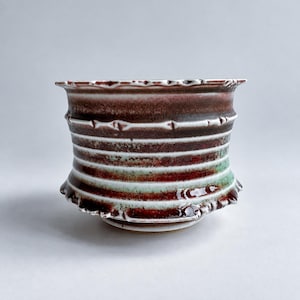 May include: A ceramic bowl featuring a flared rim and a tapered base, decorated with horizontal ridges and a glaze that transitions between deep maroon, dark brown, and soft mint green, with white accents highlighting the textured surface.