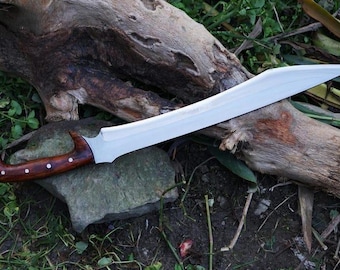 May include: A curved, silver-colored blade with a wooden handle. The handle is dark brown with white accents. The blade is resting on a rock and wood, with green foliage in the background. The sword is a handmade item.
