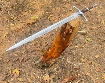 May include: A long, silver sword with a cross-shaped hilt and a brown leather-wrapped handle. The sword is resting on a weathered wooden log, surrounded by fallen leaves and foliage.