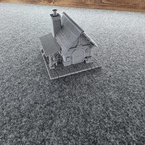 May include: A miniature gray model of a house with a chimney, a sloped roof, and a small porch. The house is made of a textured material and sits on a brick-like base. The background is a gray surface.