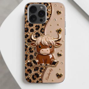 May include: A phone case featuring a brown cartoon bull with horns, centered on a split background of gold leopard print and solid beige. The design includes small heart-shaped accents, scattered rhinestones, and the name Kimberly in script.