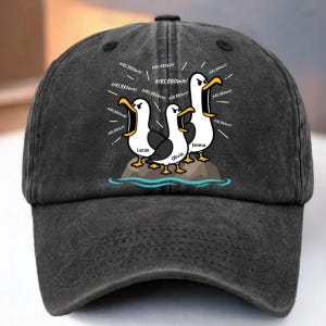 Peut inclure: Une casquette de baseball gris anthracite ornée d'un motif représentant trois mouettes sur un rocher. Les oiseaux ont le bec ouvert et le texte "MRS. BROWN!" est répété autour d'eux. Les prénoms "Lucas", "Olivia" et "Emma" sont inscrits.