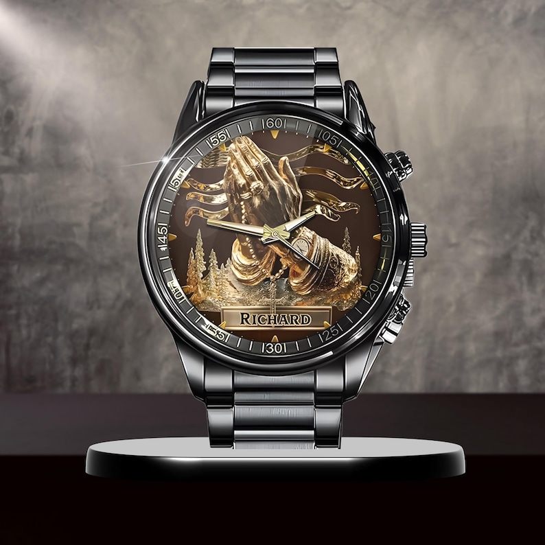 Puede incluir: Un reloj de pulsera negro con un dise&ntilde;o dorado detallado en la esfera, que presenta manos rezando y el nombre "RICHARD". El reloj tiene una correa de metal negro y una esfera redonda con marcadores de tiempo.