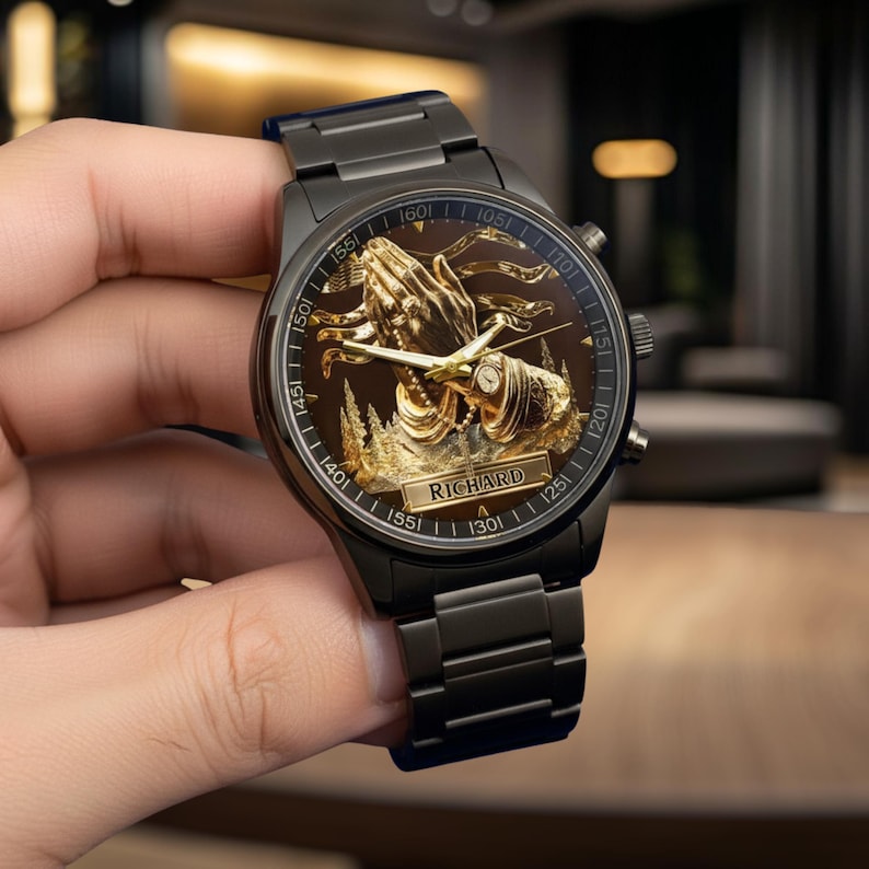 Puede incluir: Reloj de pulsera negro con un dise&ntilde;o dorado de manos rezando en la esfera. El reloj tiene una correa de metal negro y un fondo marr&oacute;n. El nombre "RICHARD" se muestra en la esfera.