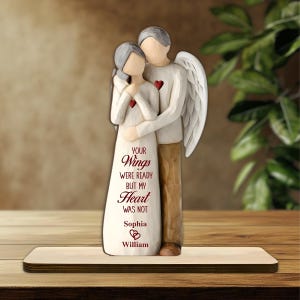 May include: A wooden figurine of a couple embracing, with one figure having angel wings. The woman wears a white dress, and the man has a red heart on his chest. The text on the dress reads "Your Wings Were Ready But My Heart Was Not".