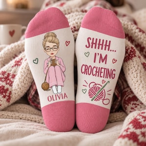 May include: A pair of white socks with pink toe and heel sections. The left sock features an illustration of a person with glasses and a bun, holding yarn, with the name OLIVIA printed below. The right sock displays the text SHHH... I'M CROCHETING.