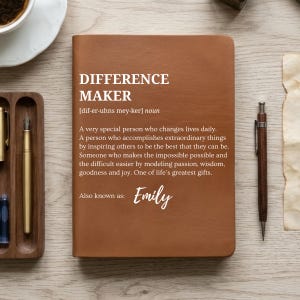 May include: A brown leather journal with the words "DIFFERENCE MAKER" and a definition printed on the cover. The journal also includes the name "Emily". A pen and a cup of coffee are on the wooden surface.