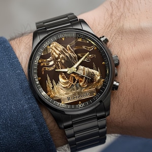 Puede incluir: Reloj de pulsera negro con un dise&ntilde;o detallado de manos rezando en color dorado sobre una esfera marr&oacute;n. El reloj tiene una correa de metal negro y el nombre "RICHARD" est&aacute; en la esfera.