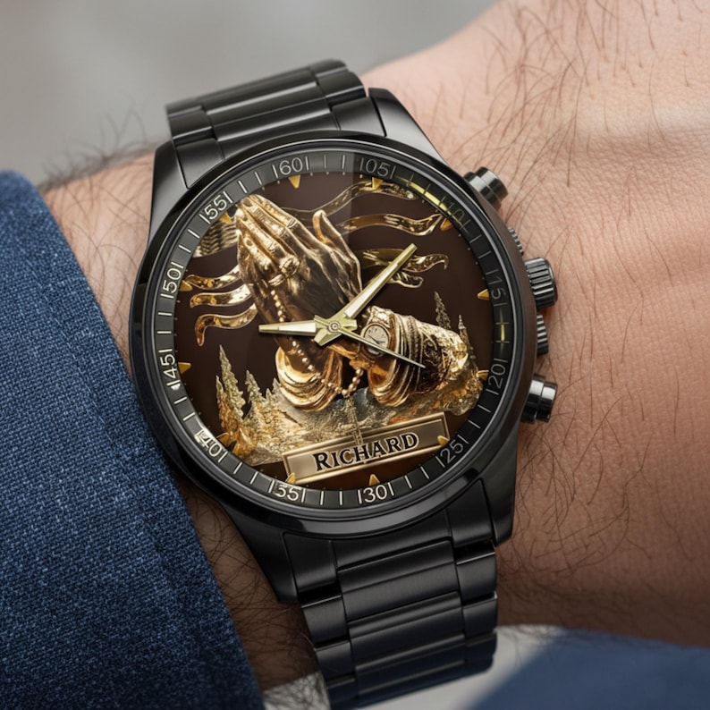 Puede incluir: Reloj de pulsera negro con un dise&ntilde;o detallado de manos rezando en color dorado sobre una esfera marr&oacute;n. El reloj tiene una correa de metal negro y el nombre "RICHARD" est&aacute; en la esfera.