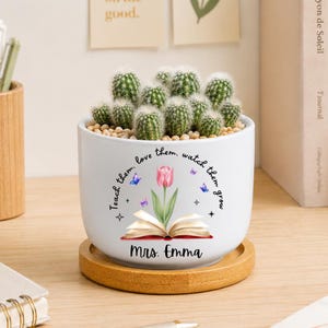 May include: A white ceramic planter containing several small green cacti, resting on a circular wooden base. The planter features a graphic of an open book with a pink tulip, blue butterflies, and the text: Teach them, love them, watch them grow, Mrs. Emma.