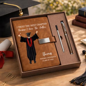 May include: A graduation gift set in a brown box. The set includes a brown leather journal with the text "I TRUST THE NEXT CHAPTER Because I know the Author" and a pen. The journal has a silver clasp and a graduation illustration.