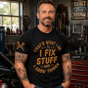 May include: A black short-sleeved t-shirt featuring orange text that reads "THAT'S WHAT I DO I FIX STUFF AND I KNOW THINGS" with a wrench graphic. The sleeve displays a crossed wrench design with the name "DANIEL" printed in the center.
