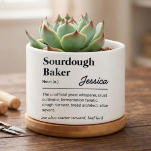 May include: A white ceramic cylindrical planter with a light green succulent plant. The planter features black text: Sourdough Baker Noun (n.) Jessica. The text includes a definition of a baker and a see also section. It sits on a round wooden base.