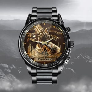 Puede incluir: Un reloj de pulsera negro con un relieve dorado detallado de manos rezando sobre una esfera marr&oacute;n. El reloj tiene una correa de metal negro y el nombre "RICHARD" est&aacute; en la esfera. El fondo muestra una cordillera.