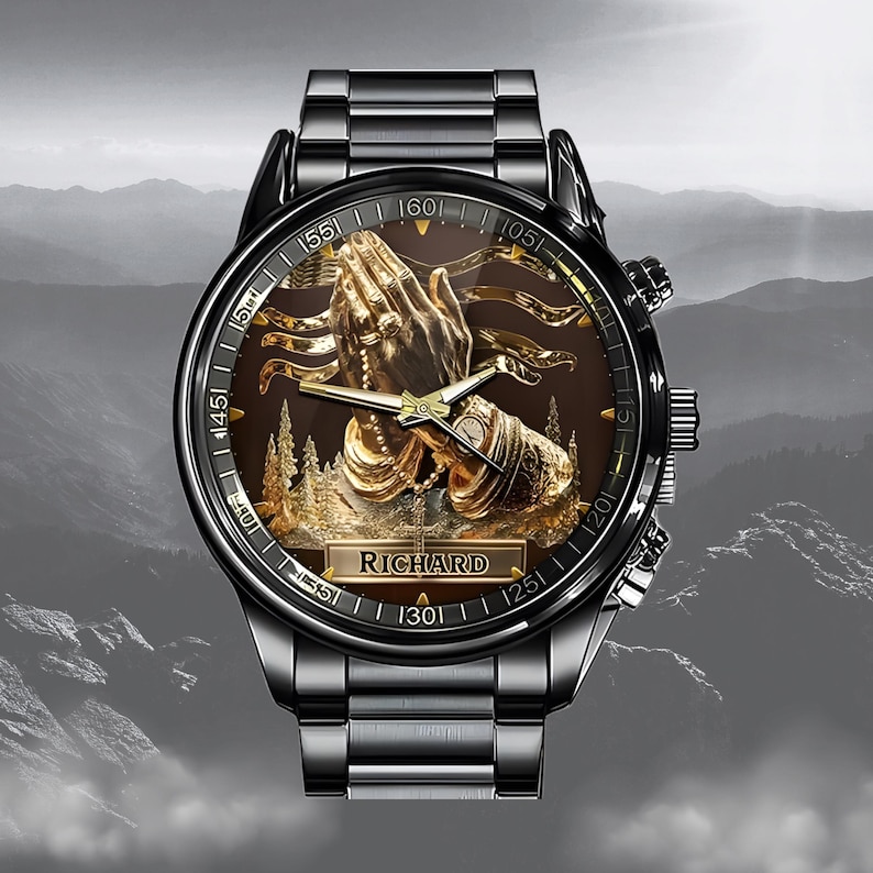 Puede incluir: Un reloj de pulsera negro con un relieve dorado detallado de manos rezando sobre una esfera marr&oacute;n. El reloj tiene una correa de metal negro y el nombre "RICHARD" est&aacute; en la esfera. El fondo muestra una cordillera.