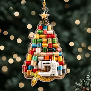 May include: A Christmas tree-shaped ornament made of stacked colorful thread spools, topped with a gold star. A white sewing machine sits at the base with the name Jennifer and the year 2026 printed on it, wrapped in a yellow measuring tape.