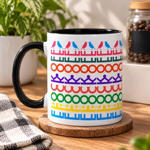 May include: A white ceramic mug with a black handle and interior, featuring a repeating horizontal pattern of pink and blue birds, followed by rows of colorful geometric shapes including circles, crowns, arches, and bars in a rainbow color palette.