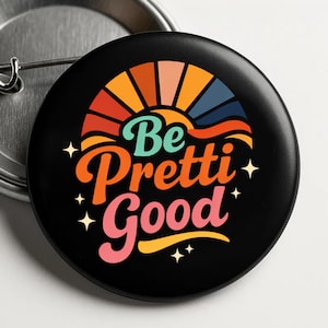 May include: A black button with the text "Be Pretti Good" in orange, pink, and teal. The design includes a retro-style rainbow and star accents. The button is round and has a pin back.