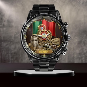 May include: Black wristwatch with a detailed face featuring the Portuguese flag, a castle, and the name "HENRY". The watch has a black metal band and a round face with gold accents.