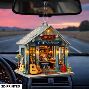 May include: A 2D printed car accessory featuring a miniature guitar shop. The shop has a blue and brown exterior with the text "JACOB'S GUITAR SHOP". Several miniature guitars are displayed outside the shop.