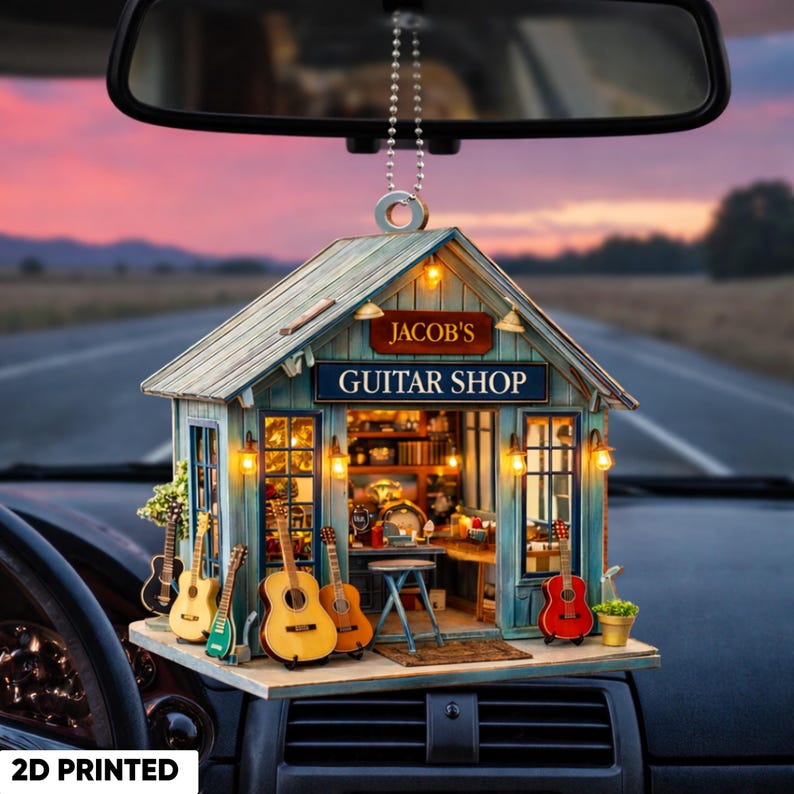 May include: A 2D printed car accessory featuring a miniature guitar shop. The shop has a blue and brown exterior with the text "JACOB'S GUITAR SHOP". Several miniature guitars are displayed outside the shop.