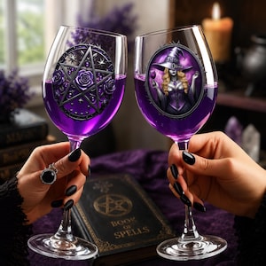 May include: Two clear wine glasses filled with purple liquid. The left glass features a black pentagram design with roses. The right glass displays an oval graphic of a witch wearing a purple hat and corset, with the name Olivia printed on the hat.