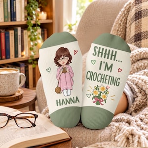 May include: A pair of off-white socks with sage green cuffs. One sock features a cartoon illustration of a person with brown hair and glasses holding yarn, with the name HANNA printed below. The other sock reads SHHH... I'M CROCHETING with a bouquet of yellow flowers.