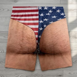 May include: Novelty shorts featuring a realistic photographic print of hairy skin on the legs and buttocks, overlaid with a graphic design of the American flag. The left side displays red and white horizontal stripes, while the right side shows blue.