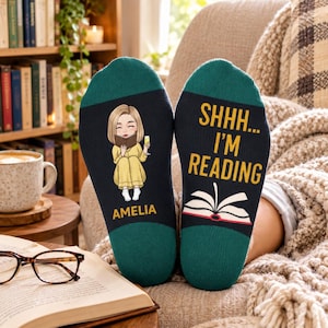 May include: A pair of dark green and black socks. One sock features a cartoon illustration of a person with blonde hair reading a book while holding a glass, with the name AMELIA printed below. The other sock displays the text SHHH... I'M READING above a graphic of an open book.