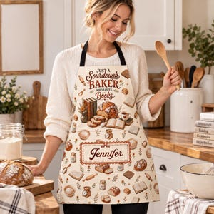 May include: A cream-colored apron featuring the text Just a Sourdough Baker Who Loves Books and the name Jennifer. The design includes illustrations of sourdough loaves, open books, rolling pins, jars, and kitchen scales in brown tones.