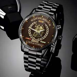 May include: Black wristwatch with a brown face featuring a gold emblem and the text "ALL GAVE SOME, 58479 GAVE ALL". The watch has a black metal band and the words "VIETNAM VETERAN" are visible on the watch face.