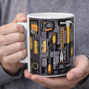 May include: White ceramic mug featuring a toolbox design with various tools, including a hammer, screwdrivers, and pliers. The tools are arranged in a dark gray background with orange and yellow accents. The mug is held in a person's hands.