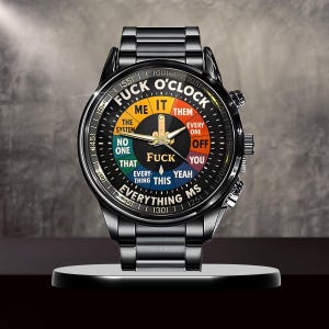May include: A black wristwatch with a unique dial design. The watch face features the text "FUCK O'CLOCK" with various phrases in a colorful segmented circle. The watch has a black metal band and a silver-colored stand.