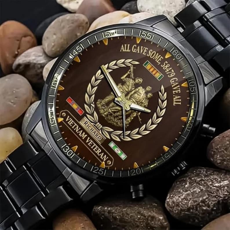 May include: Black wristwatch with a brown face featuring a gold emblem and the text "ALL GAVE SOME, 58479 GAVE ALL" and "VIETNAM VETERAN". The watch has a black metal band and is set against a background of small stones.