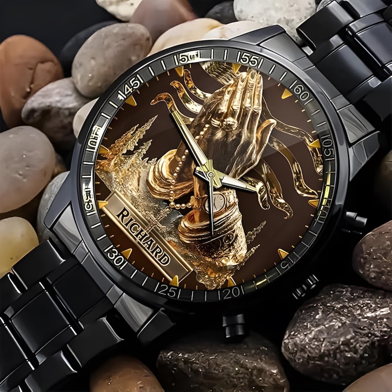 Puede incluir: Reloj de pulsera negro con esfera marr&oacute;n que presenta un dise&ntilde;o de manos rezando en color dorado. El reloj tiene una correa negra y el nombre "RICHARD" aparece en la esfera. El reloj est&aacute; sobre un fondo de peque&ntilde;as piedras.